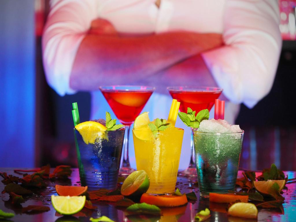 A colorful assortment of cocktails on a bar counter, garnished with citrus slices and mint leaves, with a person in a white shirt standing in the background with arms crossed.