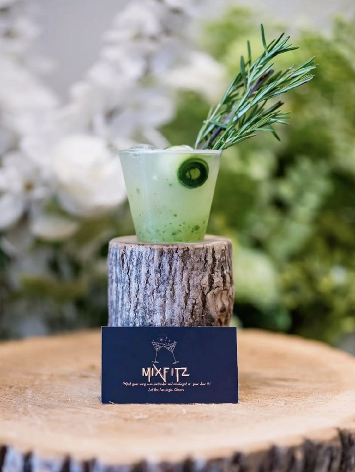 A green cocktail garnished with rosemary and a cucumber slice, served in a clear glass on a tree stump, with a MixFitz business card placed in front.